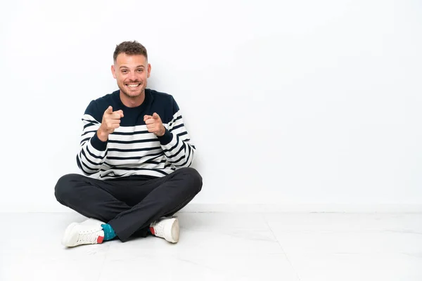 Mladý Muž Sedící Podlaze Izolovaný Bílém Pozadí Ukazuje Dopředu Usmívá — Stock fotografie