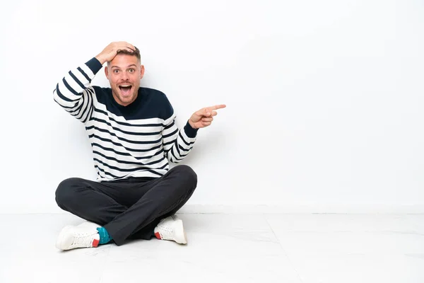 Mladý Muž Sedí Podlaze Izolované Bílém Pozadí Překvapen Ukazuje Prstem — Stock fotografie