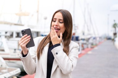 Dışarıda cep telefonu kullanan genç ve güzel bir kadın. Şaşırmış ve şok olmuş bir yüz ifadesi var.