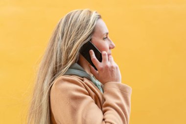 Açık havada genç sarışın bir kadın cep telefonuyla biriyle sohbet ediyor.