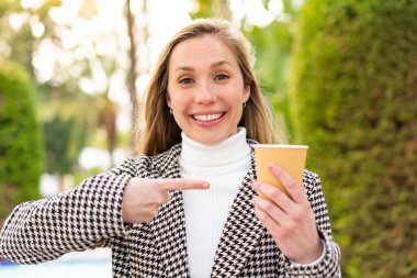 Sarışın genç bir kadın açık havada kahve tutuyor ve yüzünde sürpriz bir ifade var.