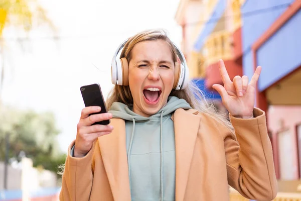 Молодая Блондинка Открытом Воздухе Слушает Музыку Помощью Мобильного Жеста Рок — стоковое фото