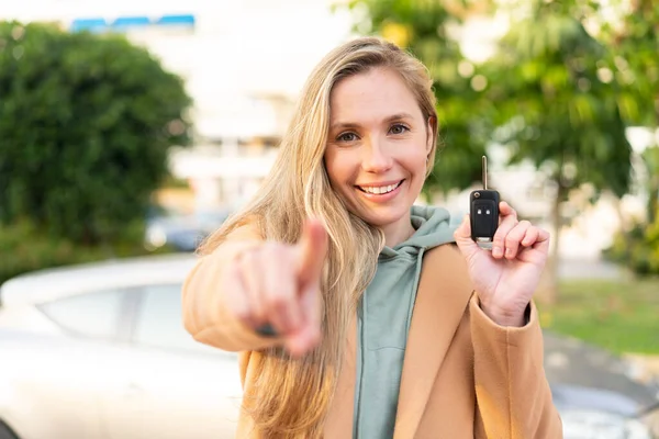 Young blonde woman holding car keys at outdoors points finger at you with a confident expression