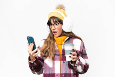 İzole edilmiş beyaz arka plan üzerinde kışlık kuklalar takan genç beyaz bir kadın elinde kahve ve bir cep telefonu tutuyor.