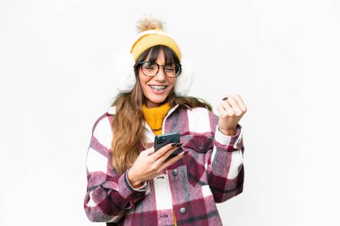 Young caucasian woman wearing winter muffs over isolated white background with phone in victory position
