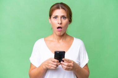 Orta yaşlı beyaz kadın izole edilmiş arka planda kameraya bakarken şaşırmış bir ifadeyle cep telefonuna bakıyor.