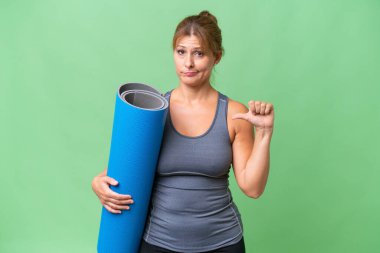 Orta yaşlı sporcu bir kadın yoga derslerine giderken elinde paspas tutarken olumsuz bir ifadeyle baş parmağını aşağı gösteriyor.