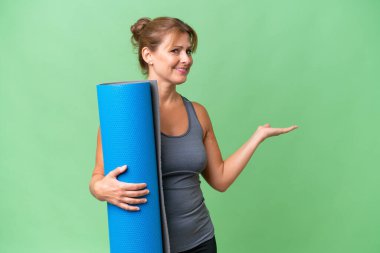 Orta yaşlı sporcu bir kadın yoga derslerine giderken izole bir arka planda paspas tutuyor ve davet etmek için ellerini yana uzatıyor.