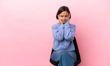 Genç melez bir kadın pembe arka planda izole edilmiş bir sandalyede oturuyor hayal kırıklığına uğramış ve kulaklarını kapatmış.