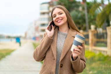 Genç, güzel, sarışın bir kadın cep telefonu kullanıyor ve elinde mutlu bir ifadeyle kahve tutuyor.