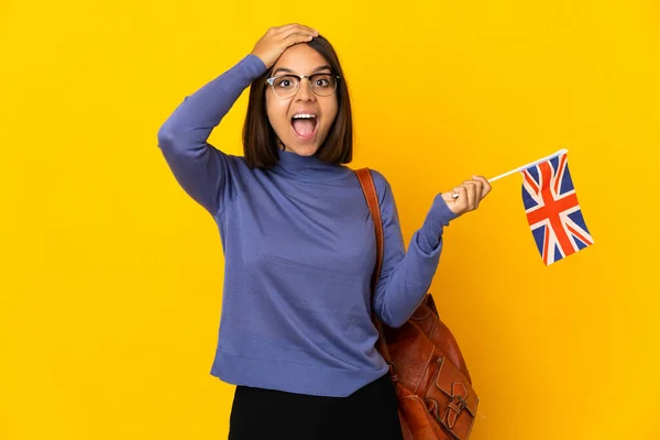 年轻的拉丁女子举着一面英国国旗 黄色背景孤立 惊讶地将手指指向侧面 — 图库照片