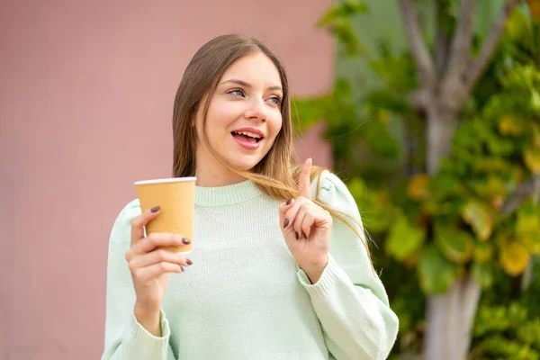 年轻漂亮的金发女人在户外拿着一杯咖啡 一边举手表决 一边想知道问题的答案 — 图库照片