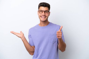 Young caucasian man isolated on white background holding copyspace imaginary on the palm to insert an ad and with thumbs up
