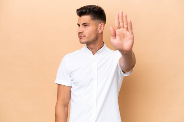 Beyazlı genç adam bej arka planda izole edilmiş. Durma hareketi yapıyor ve hayal kırıklığına uğruyor.