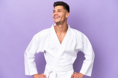 Mor arka planda karate yapan genç beyaz adam kolları kalçasında ve gülümsüyor.