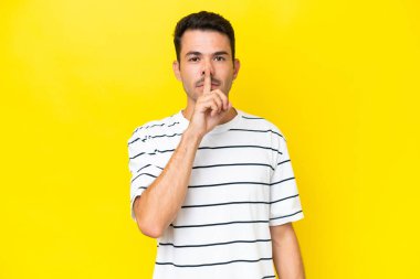 Soyutlanmış sarı arka planda genç yakışıklı bir adam sessizlik işareti gösteriyor. Parmağını ağzına sokuyor.