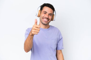 Soyutlanmış beyaz arka planda genç yakışıklı adam müzik dinliyor ve başparmağını kaldırıyor.