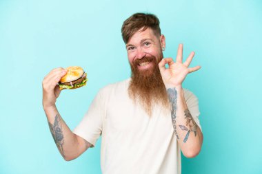Uzun sakallı kızıl saçlı bir adam mavi arka planda izole edilmiş bir burger tutuyor. Parmaklarıyla tamam işareti gösteriyor.
