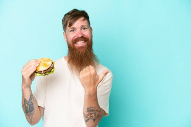 Uzun sakallı, elinde mavi arka planda izole edilmiş bir burger tutan kızıl saçlı adam bir ürünü sunmak için kenarı işaret ediyor.