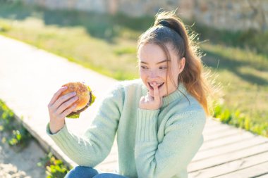 Genç, güzel bir kız, elinde sürpriz ve şok olmuş bir yüz ifadesiyle dışarıda hamburger tutuyor.
