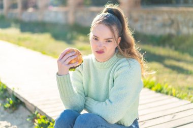 Genç ve güzel bir kız elinde burgerle dışarıda üzgün bir ifadeyle.