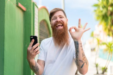 Kızıl saçlı sakallı adam dışarıda cep telefonu kullanıyor. Parmaklarıyla işaret veriyor.