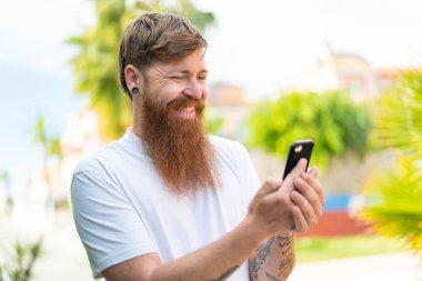 Sakallı kızıl saçlı adam cep telefonuyla mesaj ya da e-posta yolluyor.