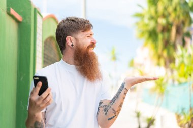 Sakallı kızıl saçlı bir adam dışarıda cep telefonu kullanıyor ve yüzünde sürpriz bir ifade var.