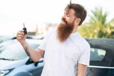 Kızıl saçlı, sakallı bir adam arabanın anahtarlarını açık havada tutuyor, mutlu bir ifadeyle.