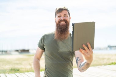 Kızıl saçlı, sakallı bir adam elinde tabletle dışarıda mutlu bir ifadeyle bekliyor.