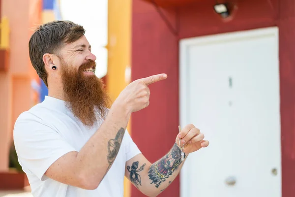 Redhead man with beard holding home keys at outdoors pointing to the side to present a product
