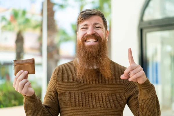 Redhead man with beard holding a wallet at outdoors pointing up a great idea