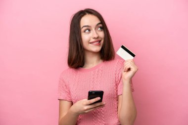 Genç Ukraynalı bir kadın pembe arka planda izole edilmiş cep telefonuyla kredi kartıyla alışveriş yapıyor.