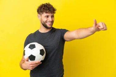 Yakışıklı genç futbolcu sarı arka planda izole edilmiş bir şekilde baş parmak hareketi yapıyor.