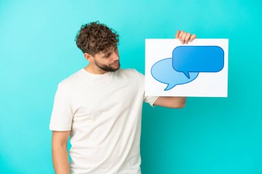 Genç beyaz adam mavi arka planda izole edilmiş. Elinde konuşma balonu ikonu olan bir levha var.