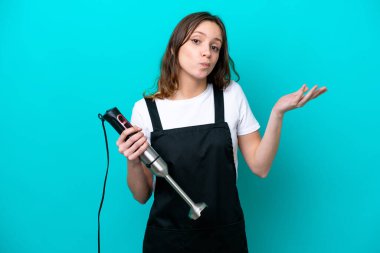 Mavi arka planda el blenderi kullanan beyaz tenli genç kadın el kaldırırken şüpheye düşüyor.