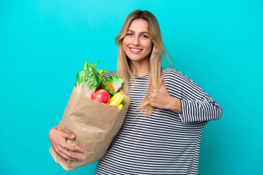 Young Uruguayan woman holding a grocery shopping bag isolated on blue background giving a thumbs up gesture