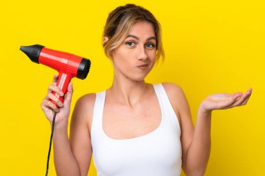 Genç Uruguaylı kadın elinde saç kurutma makinesiyle Don sarı arka planı izole ediyor. Omuzlarını kaldırırken şüphe uyandırıyor.