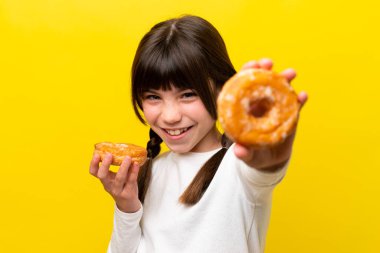 Sarı arka planda izole edilmiş küçük beyaz kız elinde mutlu bir ifadeyle donut tutuyor.