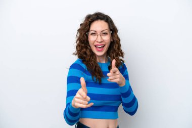 Beyaz arka planda izole edilmiş genç beyaz kadın ön tarafı işaret ediyor ve gülümsüyor.