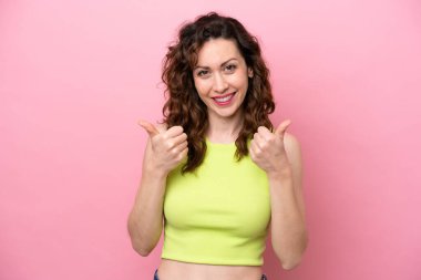 Pembe arka planda soyutlanmış genç beyaz bir kadın. Baş parmakları havada ve gülümsüyor.