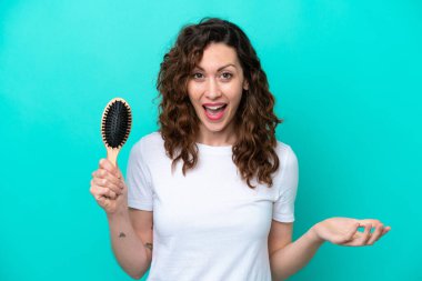Mavi arka planda saç fırçası tutan beyaz kadın şok olmuş yüz ifadesi ile izole edilmiş.