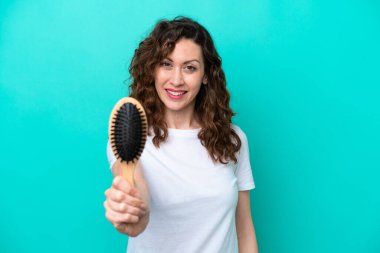 Mavi arka planda saç fırçası tutan beyaz kadın mutlu bir ifadeyle izole edilmiş.