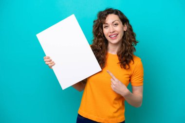 Kafkasyalı genç bir kadın mavi arka planda izole edilmiş elinde mutlu bir ifadeyle boş bir levha tutuyor ve onu işaret ediyor.