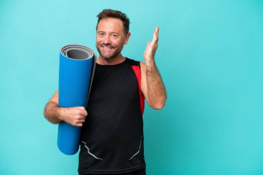 Orta yaşlı sporcu yoga derslerine giderken mavi arka planda izole edilmiş bir minder tutuyor ve gelmesi için ellerini yana uzatıyor.