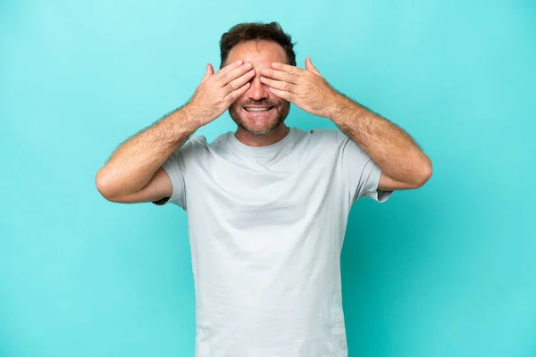 Middle age caucasian man isolated on blue background covering eyes by hands and smiling