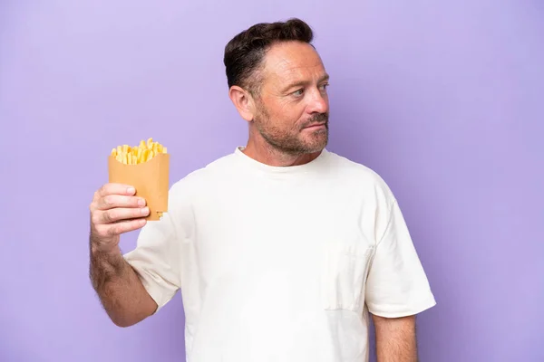 stock image Middle age caucasian man holding fried chips isolated on purple bakcground looking to the side