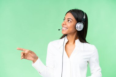 Tele pazarlamacı Afro-Amerikalı kadın izole edilmiş krom anahtar arka planda çalışan bir kulaklıkla parmak göstererek bir ürün sunuyor.