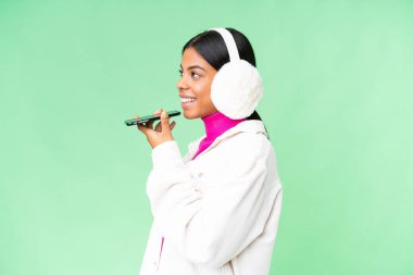 Genç Afro-Amerikalı kadın, izole edilmiş krom anahtar arka plan üzerinde kışlık manşetler takıyor ve cep telefonuyla sohbet ediyor.
