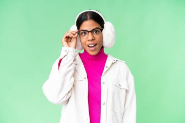 Young African American woman wearing winter muffs over isolated chroma key background with glasses and surprised